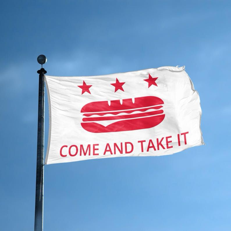 a white flag with a red sub sandwich. underneath are the words “come and take it”. above are three stars. it flies proudly against the blue sky.