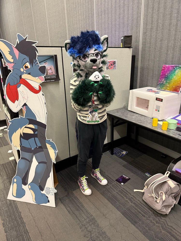 A hyena fursuiter holding a onigiri prop, standing next to a microwave and a cardboard cutout of the FurCon mascot