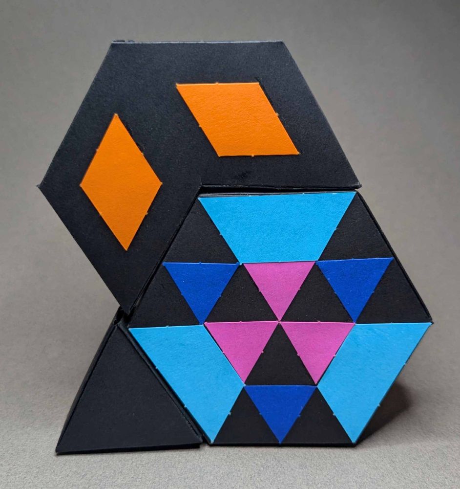 a regular hexagonal prism, an irregular hexagonal prism. and a triangular prism, all made with black paper. There are designs on the faces of the prisms that illuminate the decomposition of the larger shape. The pieces stay together by matching up magnetized surfaces. 