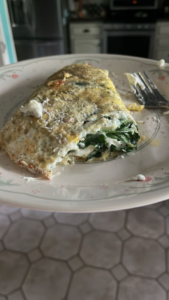 Spinach and goat cheese omelette with a bite taken out sitting on a plate with floral patterns on the edges. A tile floor is underneath and an oven and fridge sits in the background 