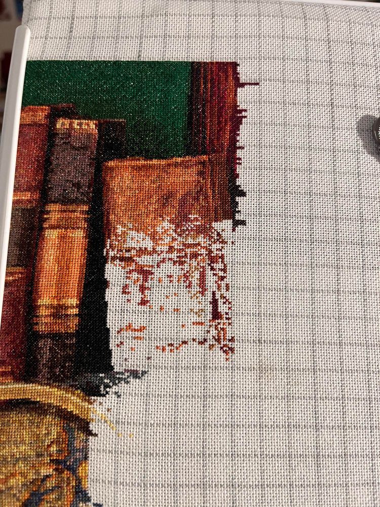White fabric with grey grid lines. There is cross stitching on the left and top in green, orange and brown in the shape of books. 