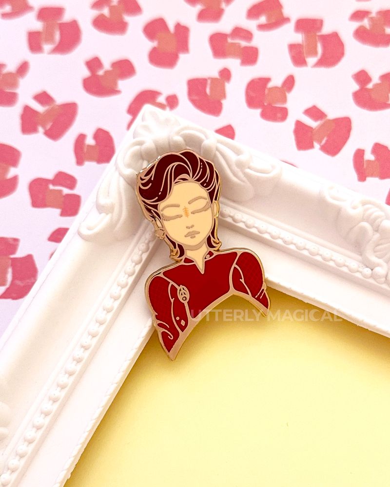 Enamel pin on a white frame on a yellow and orange background. 