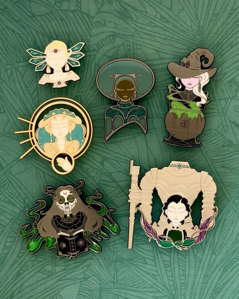 Six enamel pins on a green leaf background. 