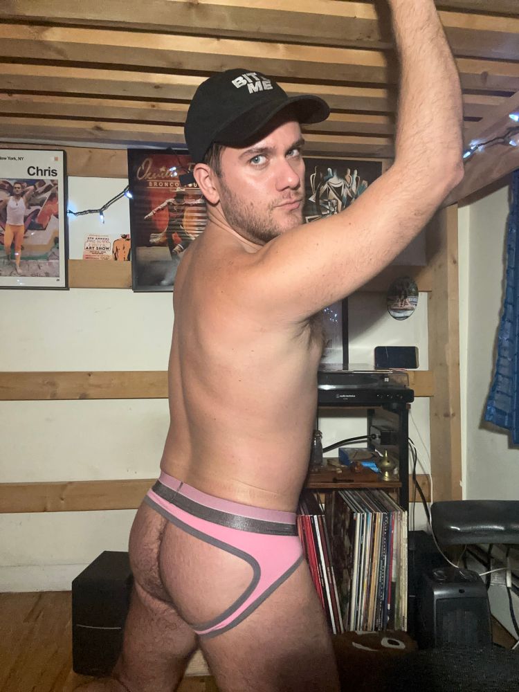 Man in a black hat (says BITE ME) and a pink jock brief poses in his bedroom. You can see posters and records. 