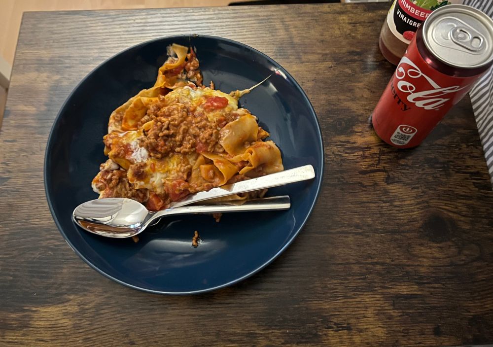 Ein Teller Lasagne auf einem kleinen Tisch mit einer Dose Cola und einer Flasche Salatdressing 