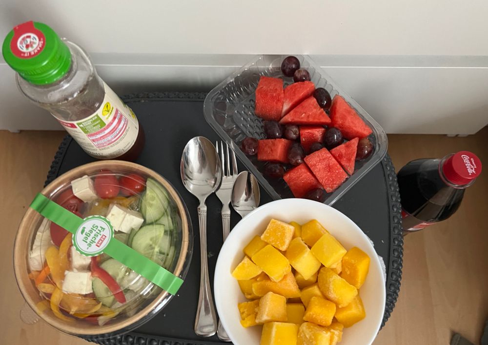 Auf einem umgedrehten Wäschekorb steht eine Flasche mit Dressing und ein Fetasalat, eine Schale mit Wassermelone und blauen Trauben und eine Schüssel mit gefrorener Mango. Zwischen Salat und Obst liegt Besteck und rechts neben dem Korb steht eine Flasche Cherry Coke
