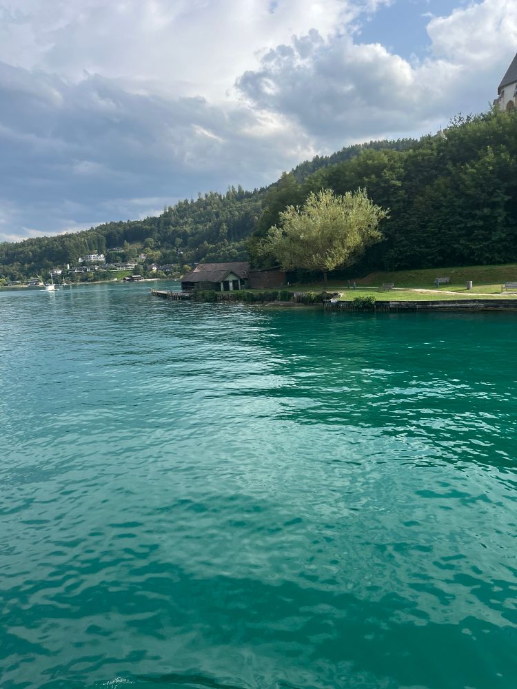 Türkises Wasser im Wörthersee mit Bootshaus und ein paar Häusern im Hintergrund 