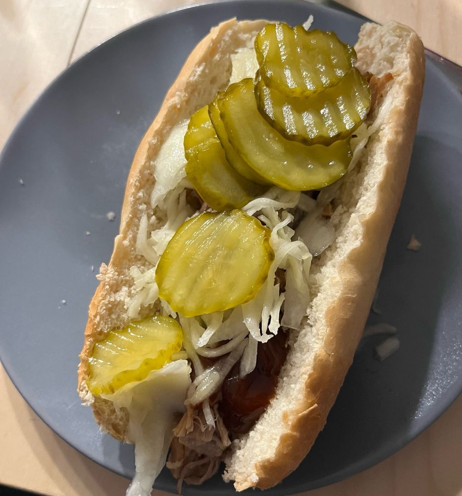 Selbst gemachtes Pull Pork versteckt sich schüchtern in einem aufgebackenen Baguette Brötchen unter einer Decke aus Barbecuesauce, Krautsalat und Burgergurken