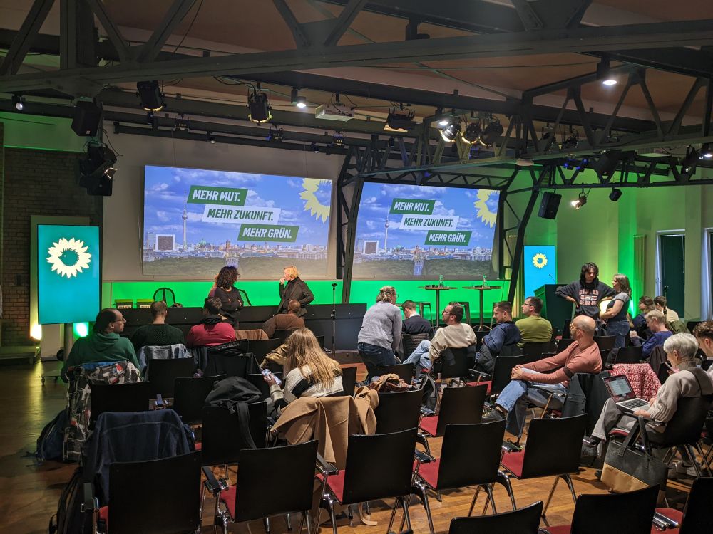 Das Bild zeigt den Raum, in dem Landesausschuss tagt. Zwei große Leinwände mit dem Slogan: "Mehr Mut. Mehr Zukunft. Mehr Grün" sind an der Wand zu sehen. Im Raum sind mehrere Stuhlreihen. Delegierte sitzen und stehen in Gruppen oder einzelnd herum.