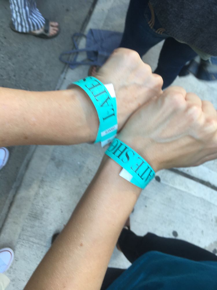 Two women hold their wrists together to show their matching green wristbands for the line up to be in the audience for an episode of Colbert’s show. The paper wristbands say “Late Show”.