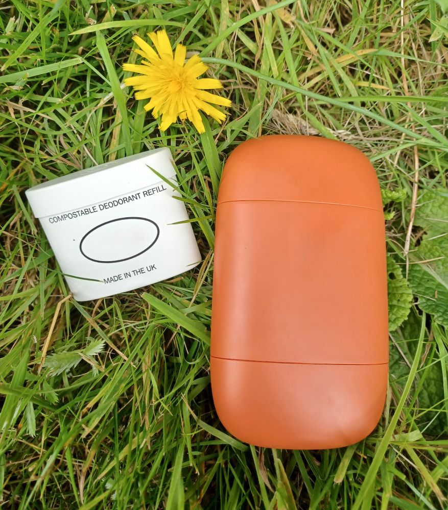 An orange refillable deodorant stick with a small white compostable deodorant refill, both laid on the grass with yellow dandelions