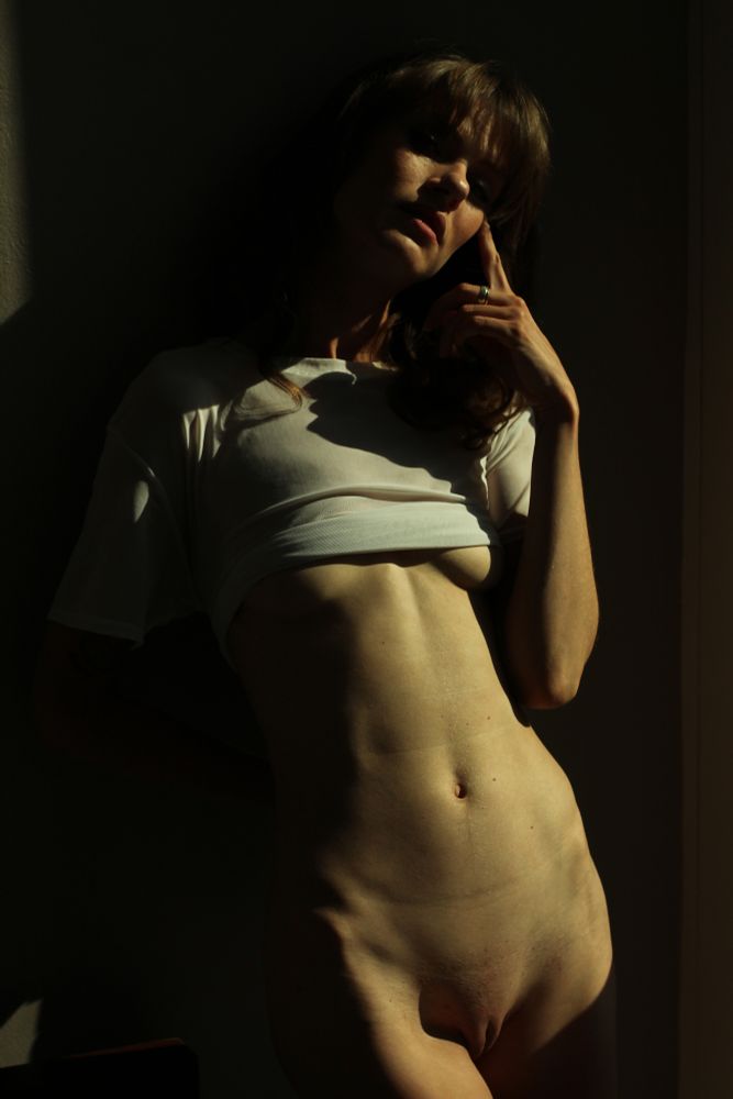 An implied nude portrait of a woman in golden hour lighting. She is wearing only a white shirt while her body is mostly covered in shadows.