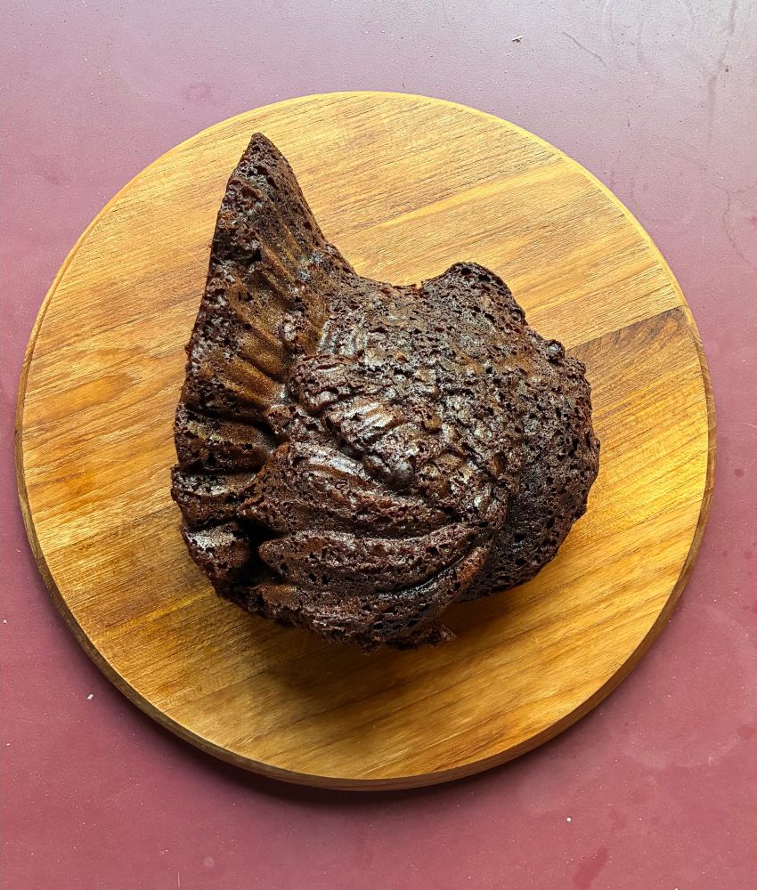 A deep brown, shiny, crumbly chocolate cake in the shape of a turkey rests on a round wooden cutting board atop a pomegranate colored table. There was not enough batter to fill the cake mould completely, resulting in a turkey without a head.