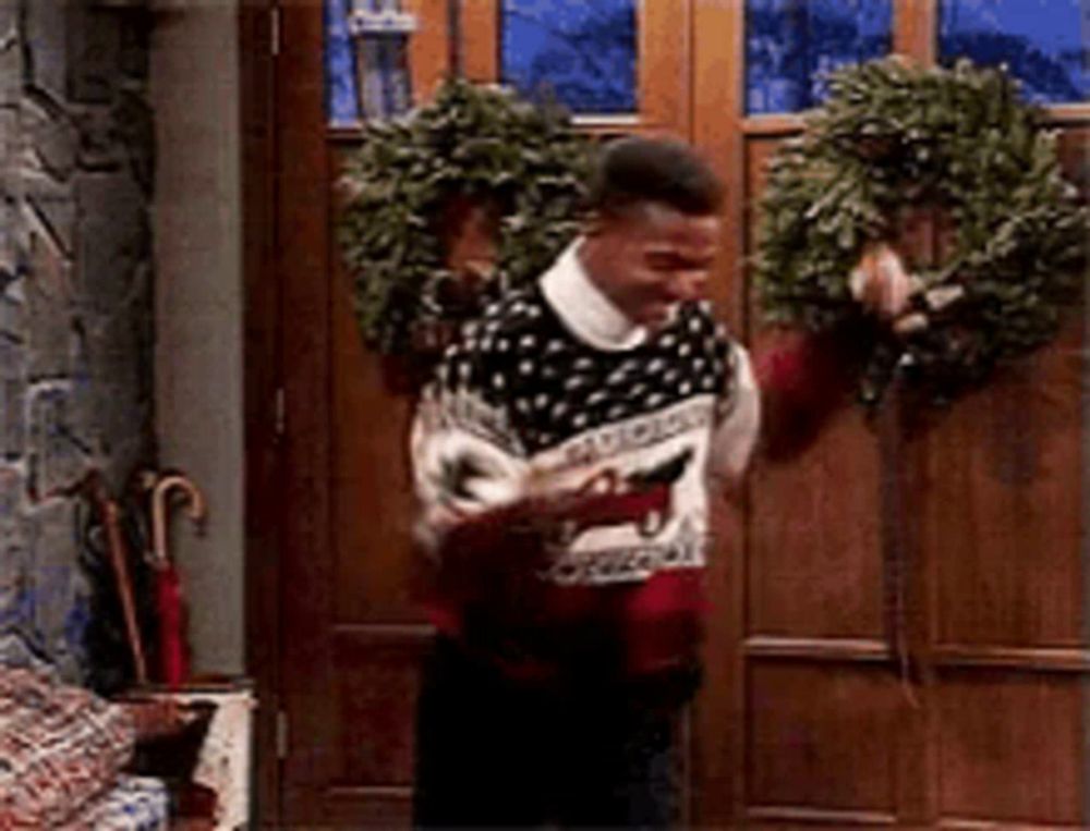 a man in an ugly christmas sweater is standing in front of a door with a wreath on it .