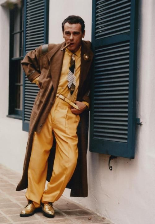 Dean Stockwell as Al from Quantum Leap. He's wearing a bright goldenrod suit (with matching gold shoes), a brown trenchcoat, and a tie that defies description. He's got a cigar in his mouth and is leaning against the side of a house with big hanging shutters.