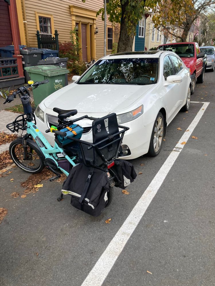 A photo of a street, with cars and a bike parked near the curb. A white car is parked within a paint outline on the asphalt, and a bike is parked in front of it. 

The white paint outline used to demarcate a spot for bicycle parking, which has now been removed.
