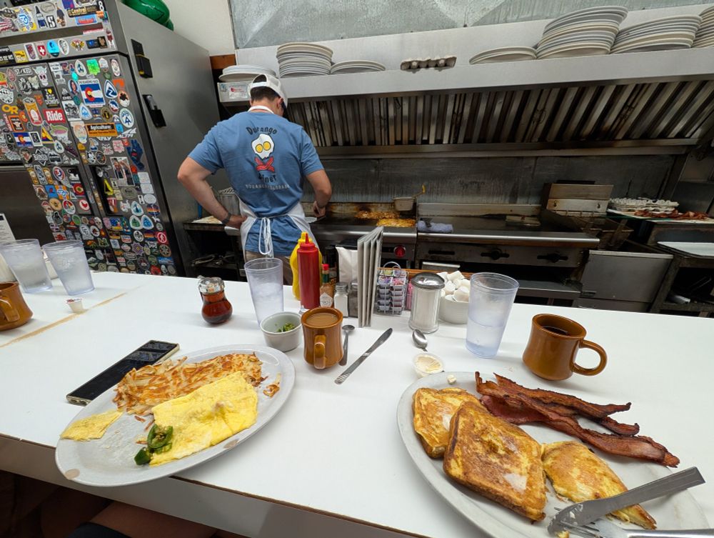 A diner counter scene where a person in a blue "Durango Diner" t-shirt cooks on a griddle behind the counter. In front of them, on the white countertop, are two plates of breakfast food: one with an omelet and hash browns, the other with French toast and bacon. Various diner condiments, drinks, and mugs are scattered around the plates. The back wall is covered in stickers.