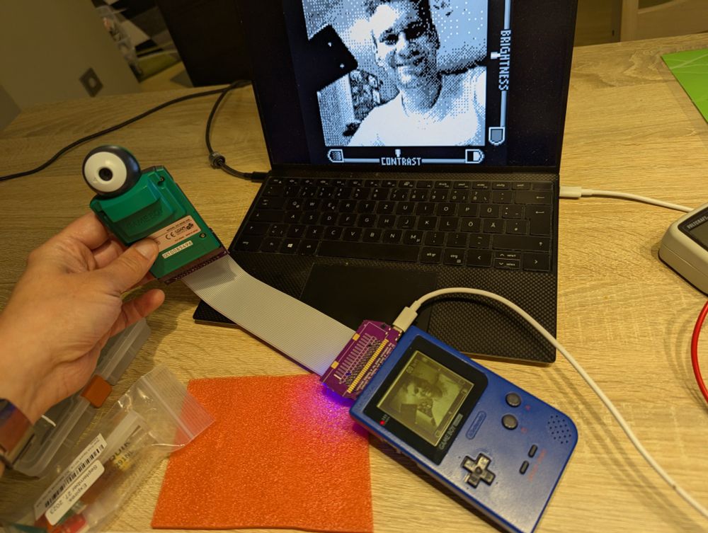 The photo shows a laptop, a Game Boy Pocket and a Game Boy Camera on a table. The Game Boy Camera is held up by a hand and connected to the Game Boy Pocket through a long ribbon cable. The top of a GB Interceptor board is sticking out from the Game Boy Pocket, which is connected to the Laptop through a USB cable. The Game Boy Pocket and the laptop both show the same image of the Game Boy Camera's view of me smiling at the camera.