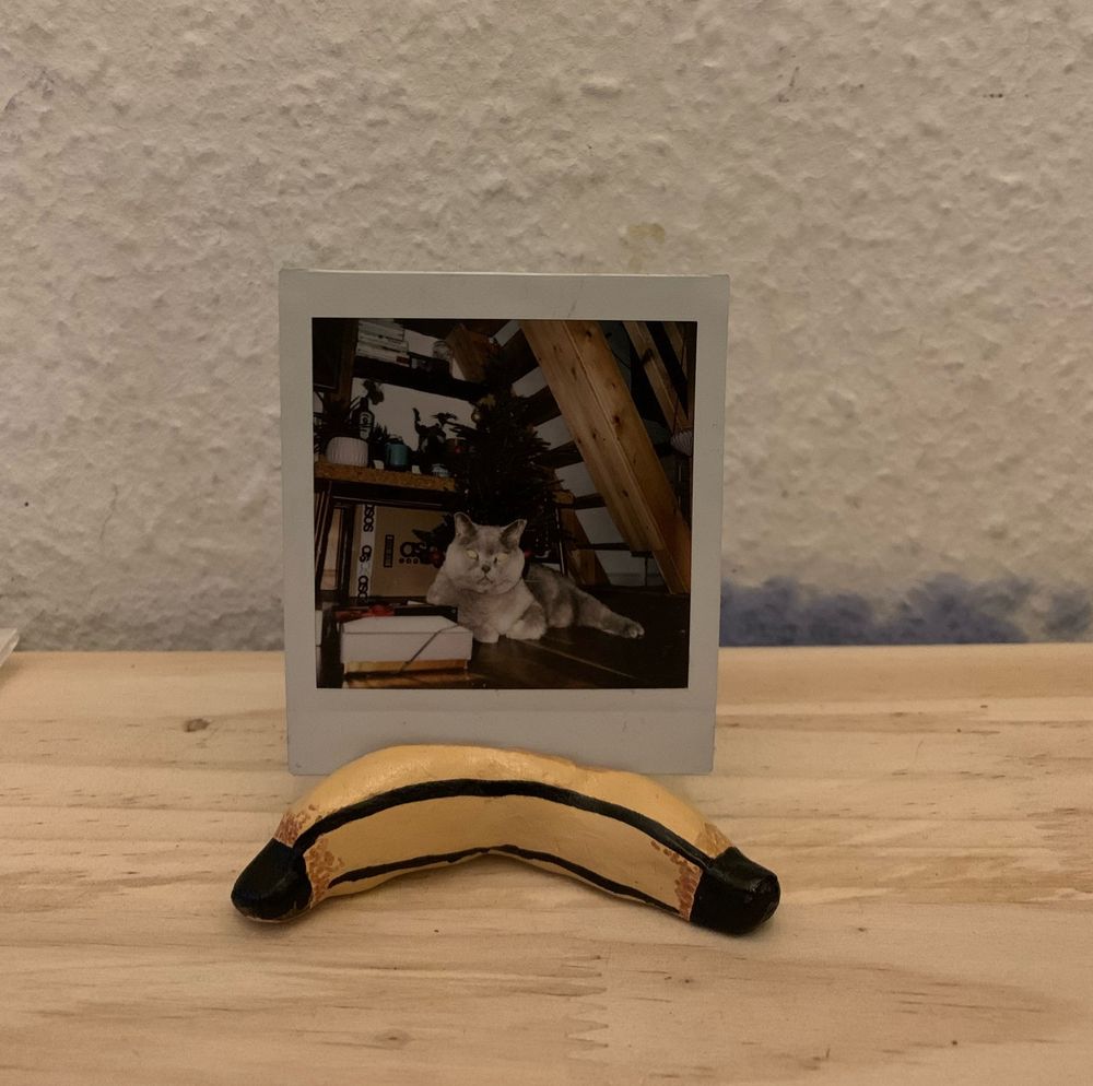 eine kleine getöpferte banane, die als polaroid halter fungiert. es steckt ein foto eines grauen katers drin, der unter einem weihnachtsbaum liegt.