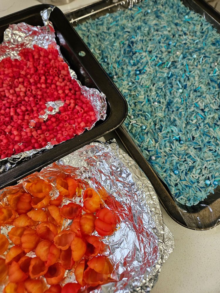 Trays of red and orange pasta and blue rice drying in my kitchen. 