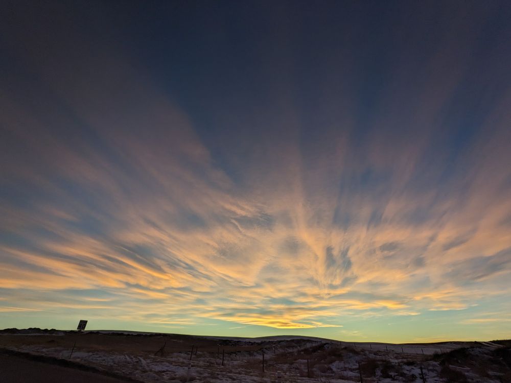 Cloud to the west at sunset. The clouds are dainty and interlaced and appear to originate from the point of sunset.  The colours shift with distance from the horizon, with bright orange and peach colours appearing low at mid-photo near the sun's position, while simultaneously dimming and shifting hues from pinks to purples to indigos and dark navy blue above and behind me.