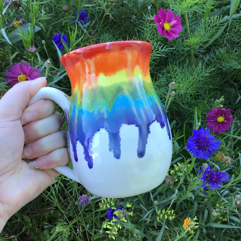 A hand made ceramic mug is being held against a backdrop of wild flowers. The mug has a rainbow colored drippy glaze. 