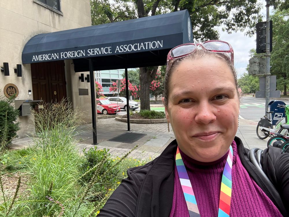 Smiling bureaucrat with a pride lanyard standing in front of the American Foreign Service Association building in Foggy Bottom, DC. 