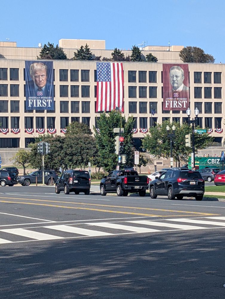 Department of Labor building in Washington DC with giant portraits of Trump and Theodore Roosevelt on either side of a giant American flag. Under each portrait are the words “American Workers First” 