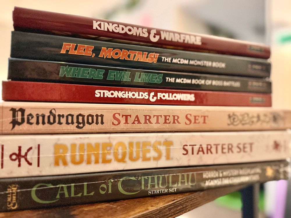 A stack of TTRPG Rulebooks and Starter sets. From top to bottom: Kingdoms & Warfare, Flee, Mortals!, Where Evil Lives, Strongholds & Followers from MCDM Productions as well as Starter Sets for Pendragon, RuneQuest, and Call of Cthulhu from Chaosium.