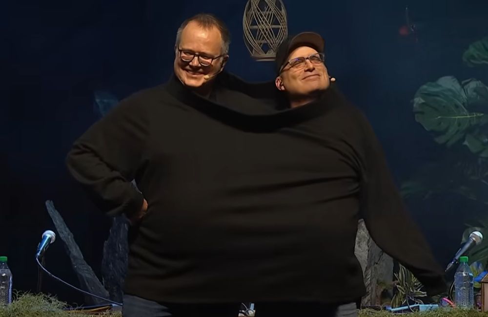 Icons of TTRPG Jeremy Crawford and Chris Perkins share a large, oversized black turtleneck sweater. They look adorably smug.