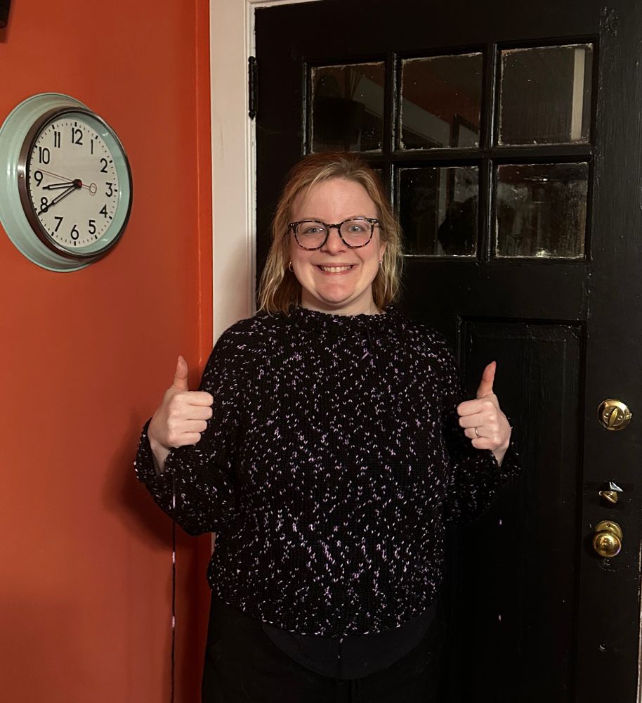A woman in a handknit black and purple speckled sweater poses with thumbs up. 