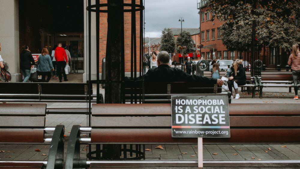 Photo in Derry of the benches outside the Foyle shopping centre. There's plenty of people in the background, and in the foreground is a sign sitting on the bench that says "homophobia is a social disease".