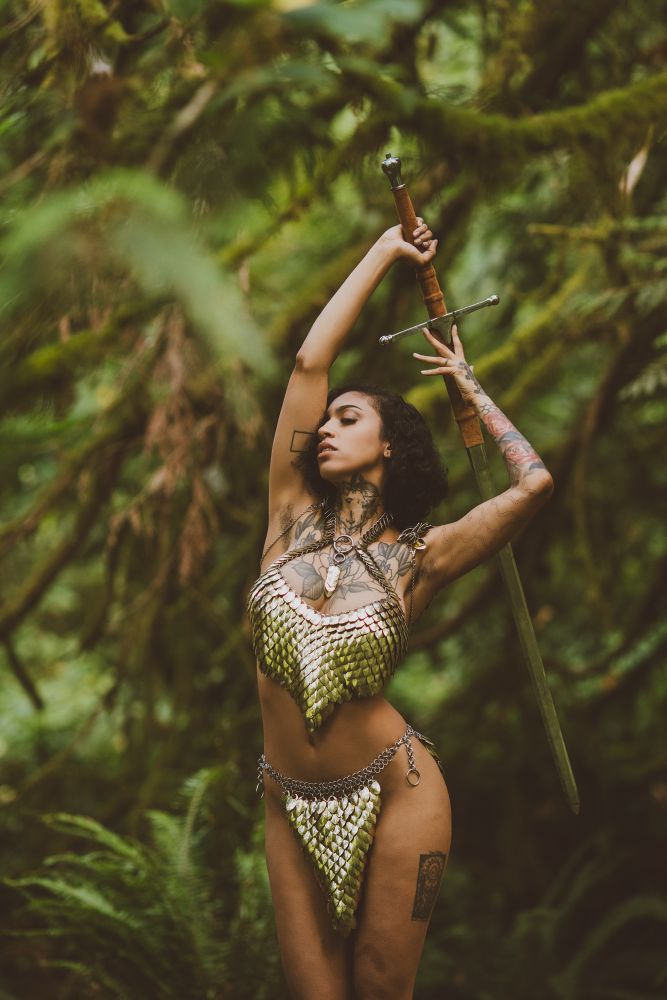 Deep in the emeral-hued forest, among the ferns and trees, where only the mist and ivy creeps, stands Ravyn Alexa. The warrior is clad in only a shining, golden chainmail. The metal catches the light, that has managed to sneak through the forest canopy, in a thousands glittering refractions that spill over her ample curves like a current of liquid gold. Her body arches up and back, forming an s-shape and putting the taught, and straining, muscles of her tummy on full display. Her arms reach over, and bend behind her head, and in her grip, a silver long sword. Her face, in profile, is slightly upturned. Light plays over her caramel skin, eyes softly closed, lashes kissing her cheeks, mouth slightly ajar, as if a steading breathe escapes her full, pink, lips.

The swords hilt is wrapped in a fawny leather and it's blade, though steel in nature, seems to be made out of forest itself. The polish on it's length being so high that it becomes camouflaged by the motley greens of the environment. The energy of the image gives the feel of a Boris Vallejo illustration given breath!