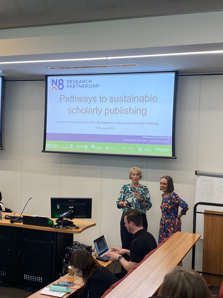 A speaker stands at the front of a lecture hall next to a large projected slide titled “Pathways to sustainable scholarly publishing”, part of the N8 Research Partnership event at the University of York on 12th June 2025. Another woman stands nearby, and attendees are seated, with some using laptops. The screen displays logos of the N8 partner universities.