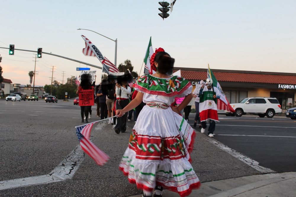 protesters crossing the street with flags from the usa, mexico and other countries. foreground is a young mexican woman with a traditional dress leading the group forward. faces are obscured.