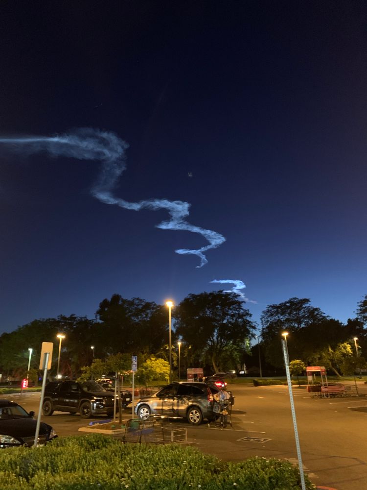spacex launch smoke swirling in the twilight sky viewed from a grocery store parking lot in southern california with trees on the horizon