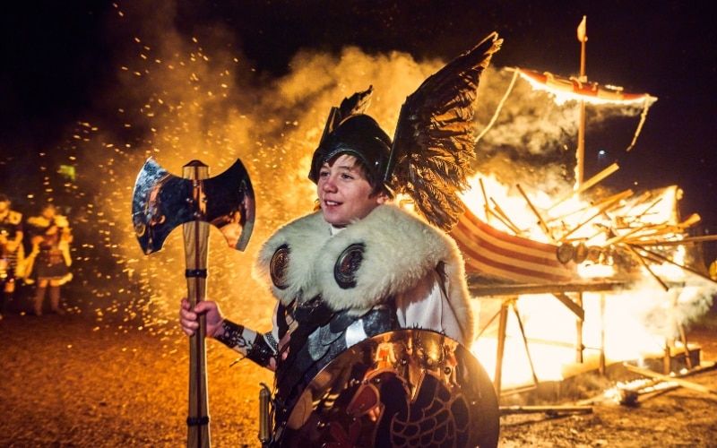 Lad dressed as a viking. He's holding an axe. There's a viking longboat in flames behind him. It looks fun