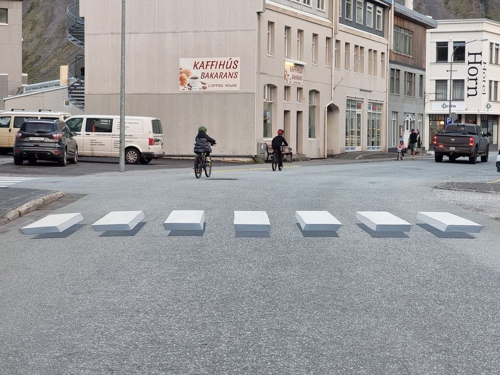 Zebra crossing in a town street, painted in such a way as to appear 3D and floating above the tarmac. Two kids are riding their bikes away from it, Kaffihús Bakarans is the cream building ahead and Horn is on the corner of the next block 