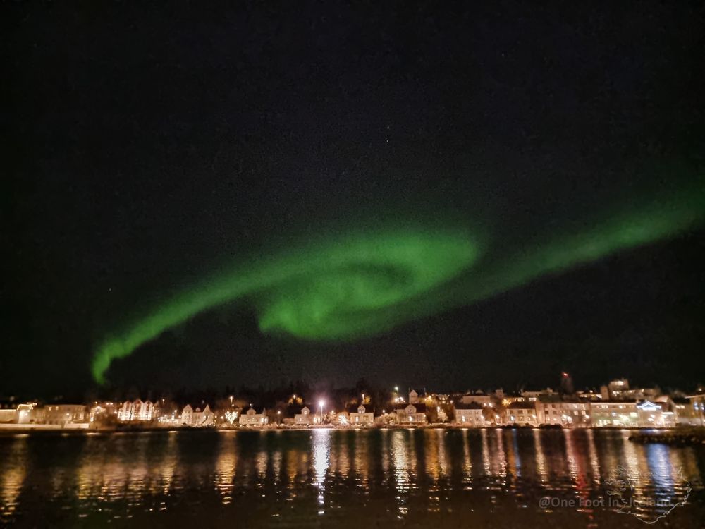 Reflection of many lit buildings around the edge of a large pond, with a spiral of green aurora in the black sky 