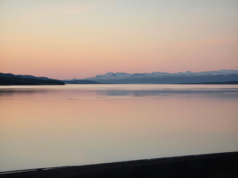 A calm lake surface with low mountains on the horizon, under a pinkish-orange sky, reflected in the water. There are one or two ripples breaking the surface, and a dark triangle at the bottom, rising from left to right, of the bridge the picture is taken from 