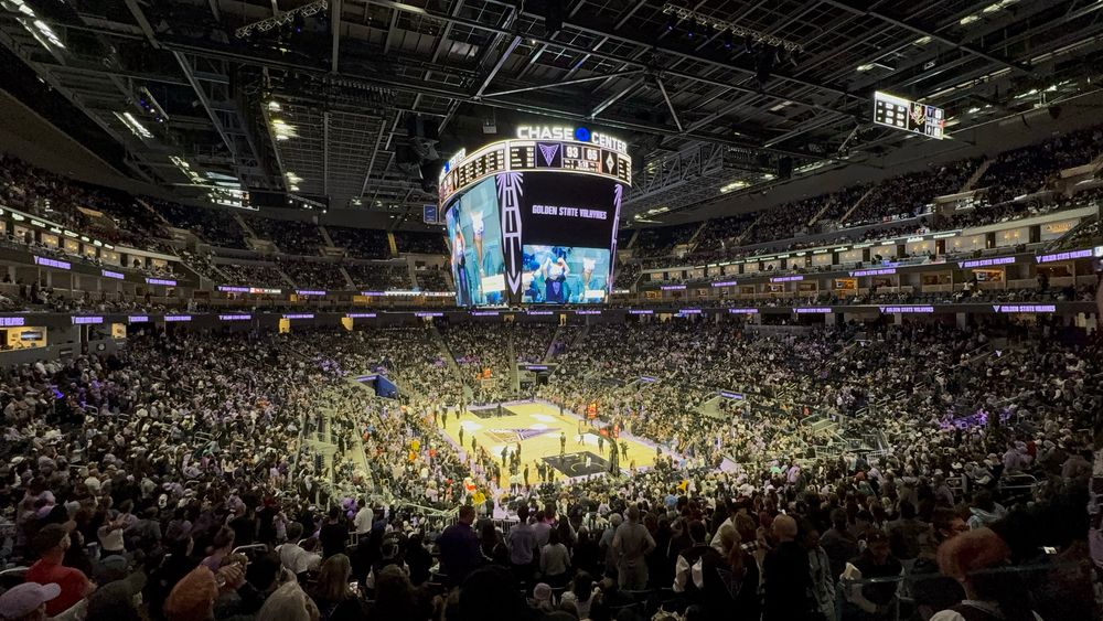 A wide-angle view of the interior of Chase Center in San Francisco during a basketball game, with fans filling the stands. The scoreboard shows the Golden State Valkyries leading 93-65. The arena’s overhead video board displays live footage of the court and spectators. 