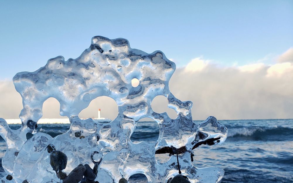 A thin sheet of lake ice has been pushed into a vertical position on the Lake Huron shoreline. It's got holes from where the pebbles were. Inside one of the holes there is a red and white painted  light beacon tower visible in the distance. 