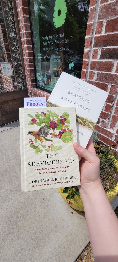 I'm holding up two books in front of a cute brick shop called Paper Hearts bookstore. The books are "Braiding Sweetgrass" and "The Serviceberry," both authored by Robin Wall Kimmerer.