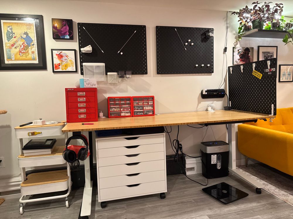 A work area showing a long desk on rising legs surrounded by black pegboards and cabinets above and below the desk.