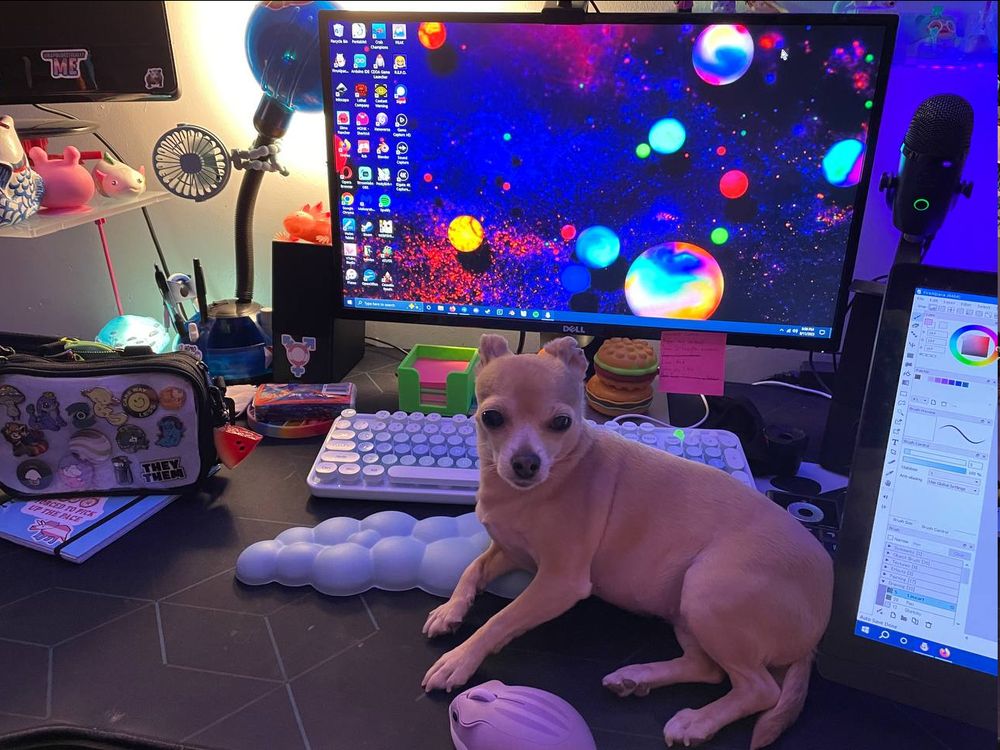 Tan chihuahua sitting on a cluttered computer desk, using a keyboard rest as a cushion