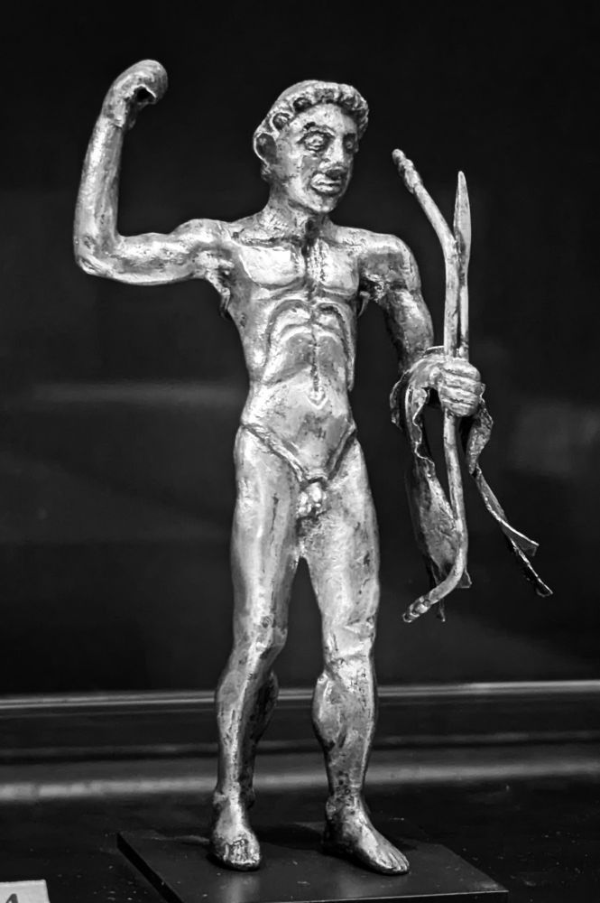 Héraclès, nu, le bras droit levé. Le bras gauche plié, tient un arc et sur son avant-bras la léonté.