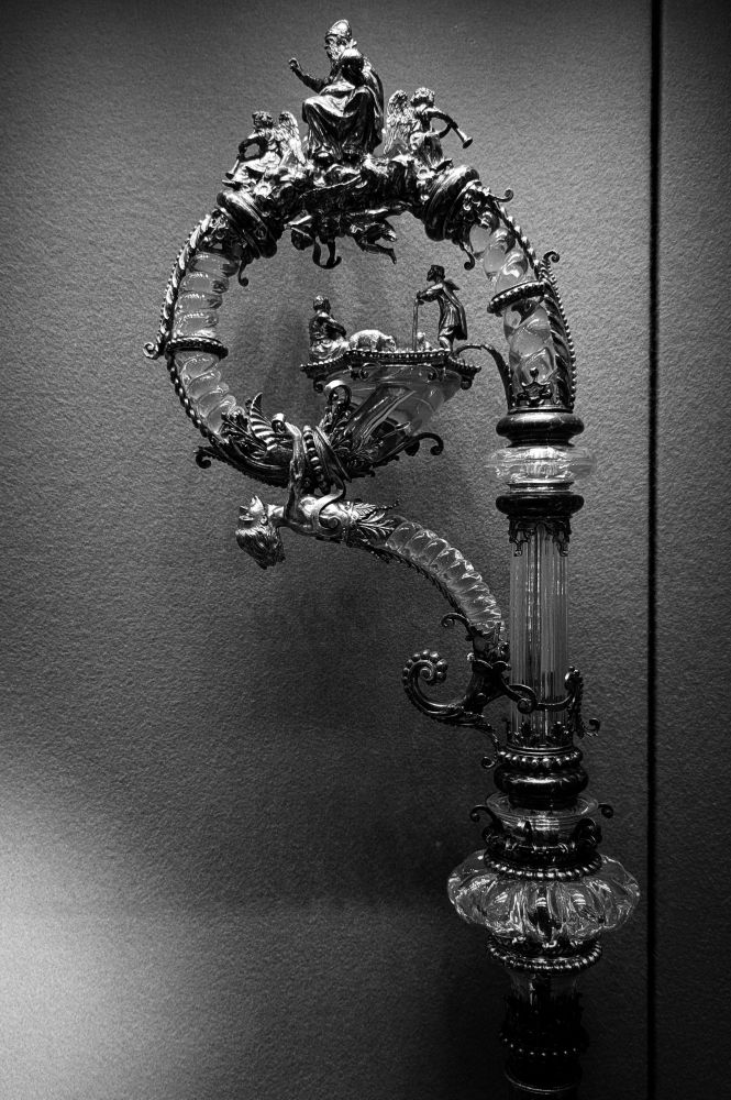 Le noeud est de forme méplate et godronnée, et se rattache au moyen de volutes d'argent doré et d'un anneau d'argent niellé à une tige cannelée, d'où naît une volute, également en cristal de roche, figurant une cordelière dont l'extrémité s'épanouit de façon à former une terrasse découpée à six pans. A la partie supérieure de la volute, que bordent des rinceaux d'argent et dont des bagues niellées ou perlées réunissent les diverses parties, on aperçoit une figure, en argent doré, du Père éternel coiffé de la tiare, bénissant avec le globe du monde, accompagné de deux figures d'anges sonnant de la trompette. Un troisième ange, tenant une banderole, se détache de la partie inférieure de ce groupe et annonce la naissance de Jésus à deux bergers figurés sur la plate-forme terminale de la volute. Cette volute est elle-même soutenue par une pièce de cristal de roche recourbée, montée en argent doré, que termine à sa partie supérieure un buste d'homme. Le bâton destiné à compléter cette crosse se compose de huit pièces cylindriques d'argent, ornées de cannelures et d'anneaux niellés.
Inscription : Gloria in excelsis Deo (sur la banderole)