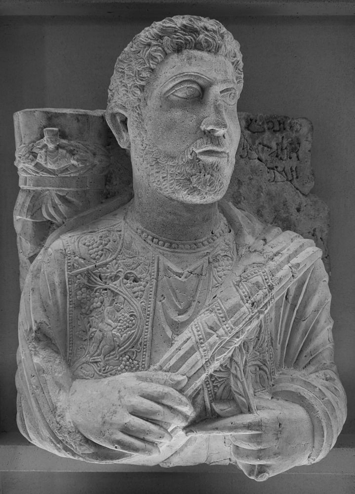 Relief funéraire : homme (buste) barbu, vêtu d'une sous-tunique bordée au cou d'un galon perlé, d'une tunique à larges bandes ornées de rinceaux de vignes et d'amours vendangeurs, et d'un manteau également pourvu d'une bande à décor végétal, paré d'une bague et tenant une palme. Au-dessus de son épaule droite est placé un modius, ceint d'une couronne de lauriers et orné d'un buste d'homme portant tunique, manteau et modius, et un voile. Inscription en palmyrénien de l'épitaphe "Yar hai fils de Elahbêl. Hélas"