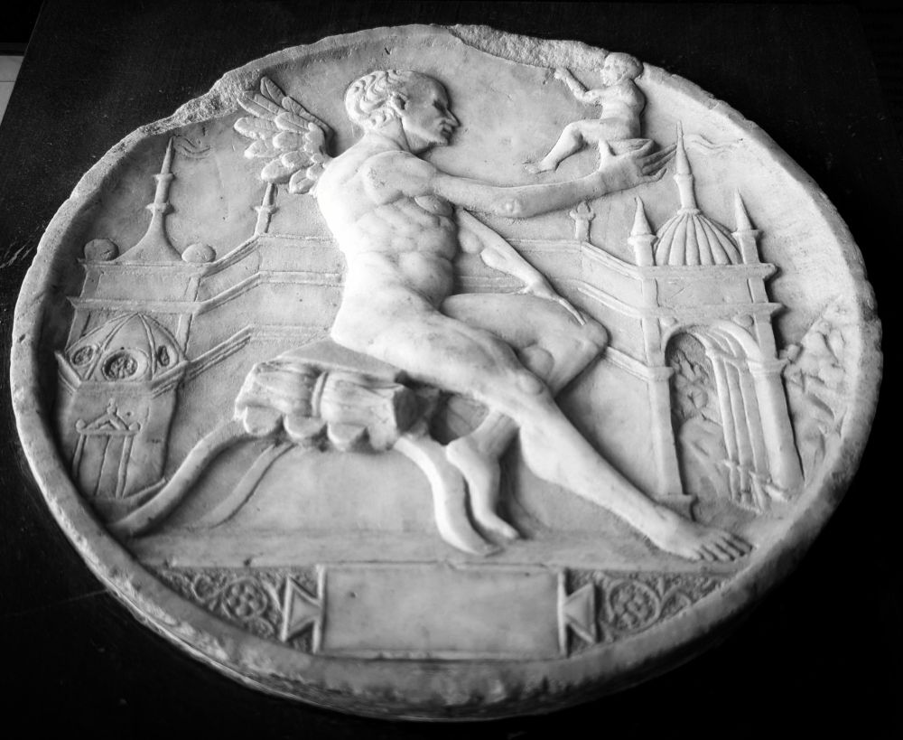Dans un décor de murs qui peut faire penser à un labyrinthe, un homme est assis, nu.
Jambe gauche repliée, jambe droite tendue, une petite paire d'ailes dans le dos, il tient à bout de bras, un jeune enfant (Icare ?) dans le creux de sa main.