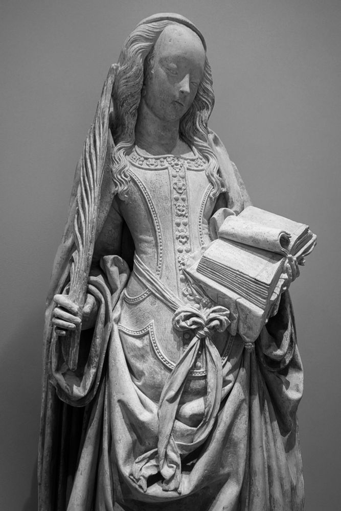 La Sainte tient dans sa main droite une palme, symbole du martyre. Dans sa main gauche un livre ouvert en deux dont une page donne l'impression d'être tournée.
La sculpture permet de montrer la finesse et les détails des différentes pièces de la robe.